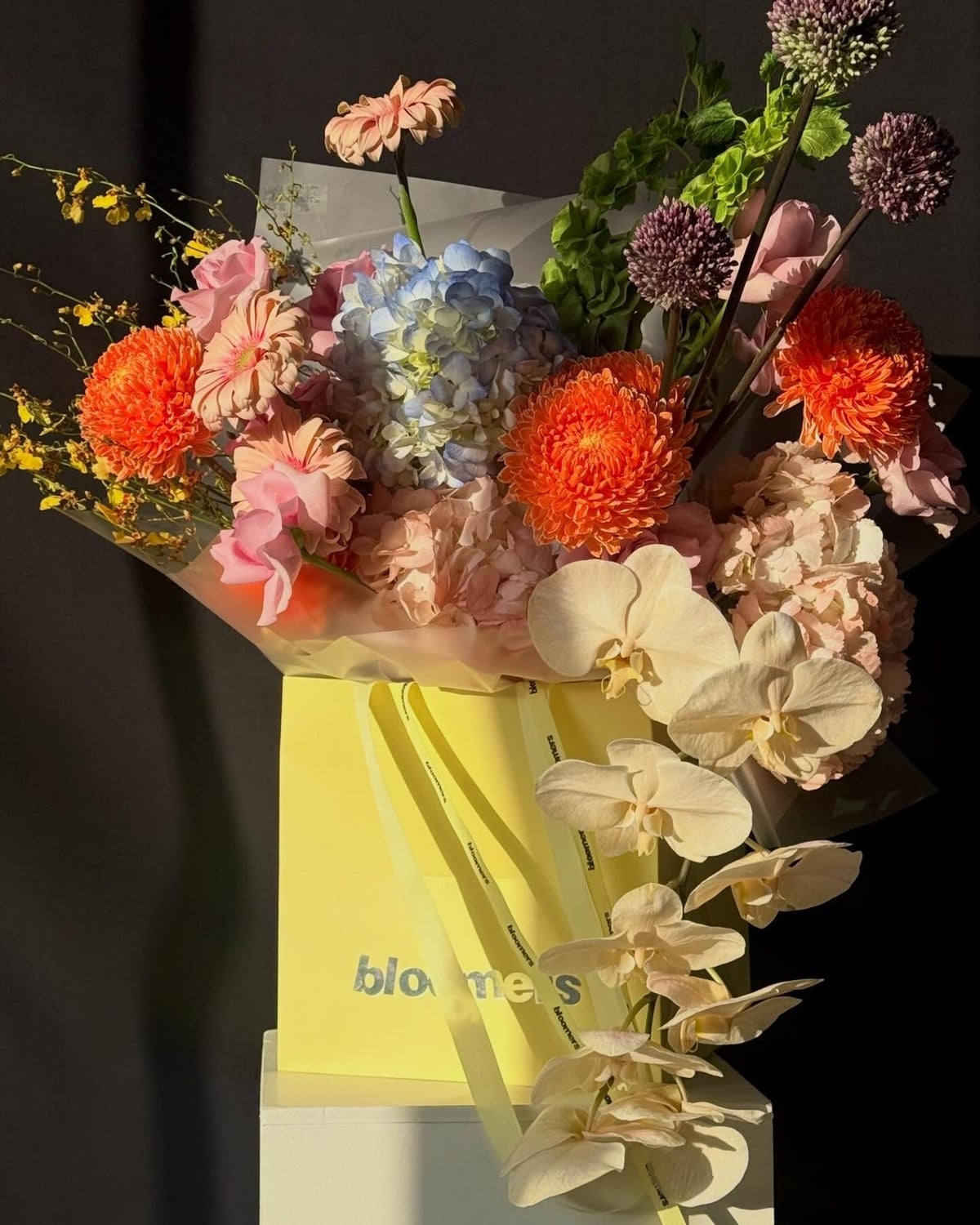 Peach, orange and pale blue flowers with cascading white orchids arranged in a yellow Bloomers bag.