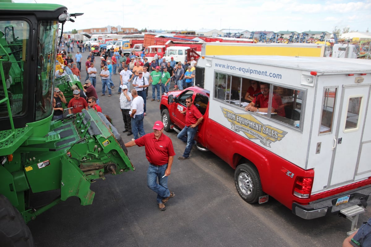 A large crowd gathers for an outdoor equipment auction. A green combine harvester sits beside a red pickup.
