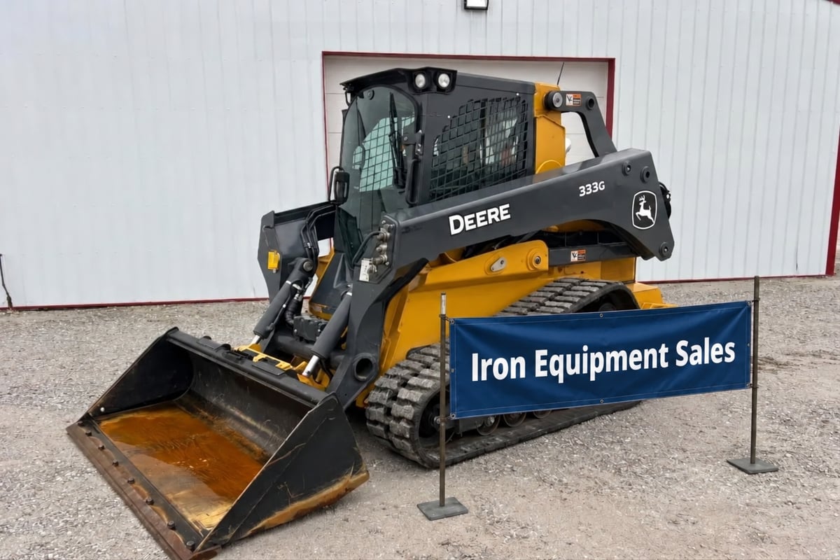 A yellow and black John Deere 333G skid steer loader with a large front bucket, parked in front of a white building. A blue b