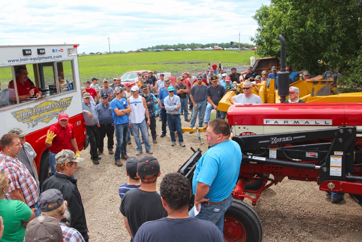 A large crowd of people gathered around a red tractor, listening to a speaker at a live auction event.